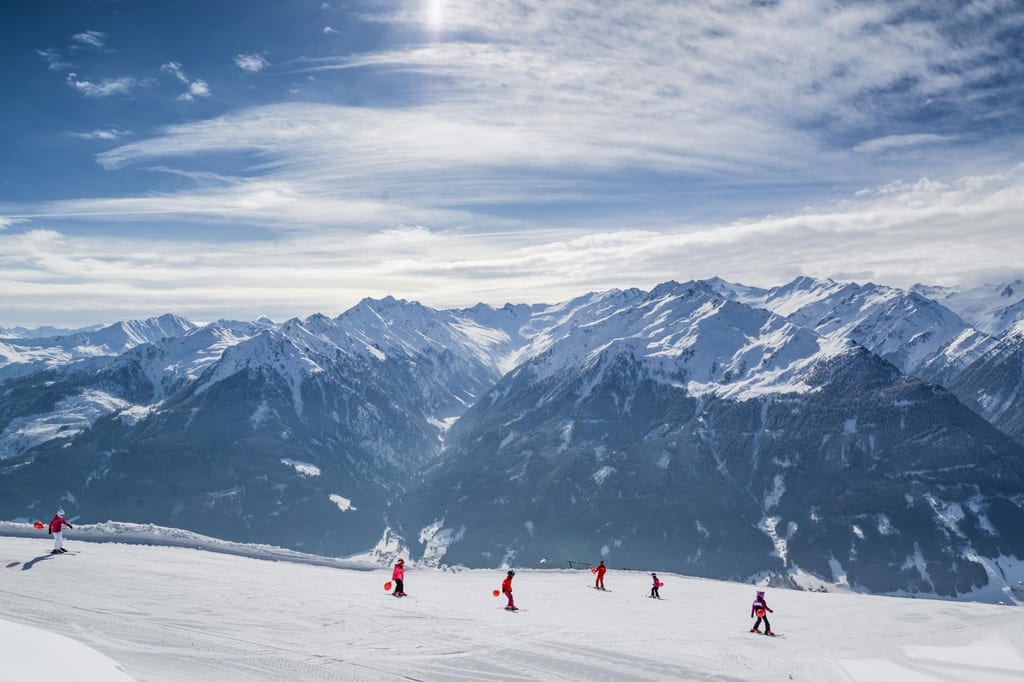 Skifahrer auf einer sonnigen Piste vor einem weiten Bergpanorama.